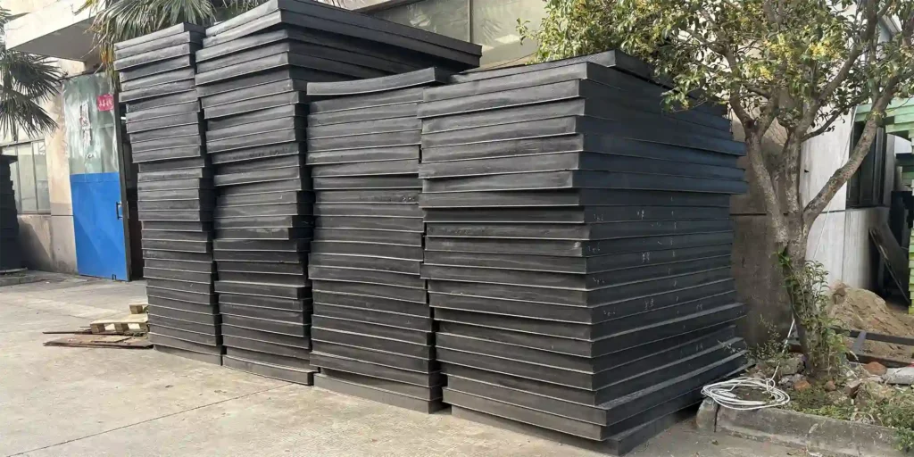 an outdoor industrial area where numerous large stacks of black plates are stored. The plates are neatly piled, and the scene includes a building with windows, palm trees, and a tree with yellow flowers. The concrete ground and some scattered materials suggest a storage or manufacturing environment for these industrial components.