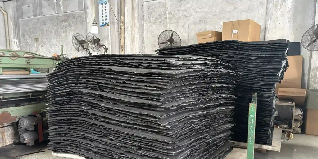 an indoor factory setting where massive stacks of black industrial sheets are stored on wheeled carts. The factory has a worn, concrete interior with old machinery, wall-mounted fans, and cardboard boxes. A safety sign on the wall reads (Working unsafely today will jeopardize your life tomorrow), emphasizing workplace safety. The scene indicates a storage or manufacturing environment for these large, black industrial components.