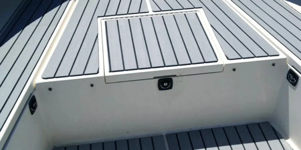 a close-up view of the deck area on what appears to be a boat or yacht. The deck is made up of horizontal grey slats, providing a textured and non-slip surface that is common in marine environments for safety purposes. In the center of the image, there’s a hatch cover with a handle, suggesting it provides access to storage space below the deck. The hatch has a white frame around it, contrasting with the grey slats of the deck. There are also visible fasteners securing the hatch cover to the deck. To the sides of the hatch, we can see the continuation of the deck extending towards the edges of the image. The overall condition of the deck looks clean and well-maintained, indicating careful upkeep of the vessel.