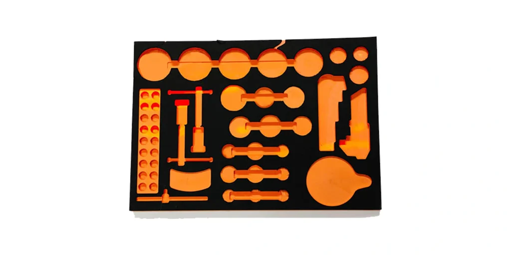 shows a rectangular, two-tone foam insert (black base with bright orange cutouts) featuring a variety of precision-machined cavities. The cutouts include circular, rectangular, and specialized shapes tailored to hold specific tools, components, or equipment securely. The contrasting colors make the cavities highly visible, emphasizing the insert’s functional, custom-fit design for organization and protection during transport or storage.