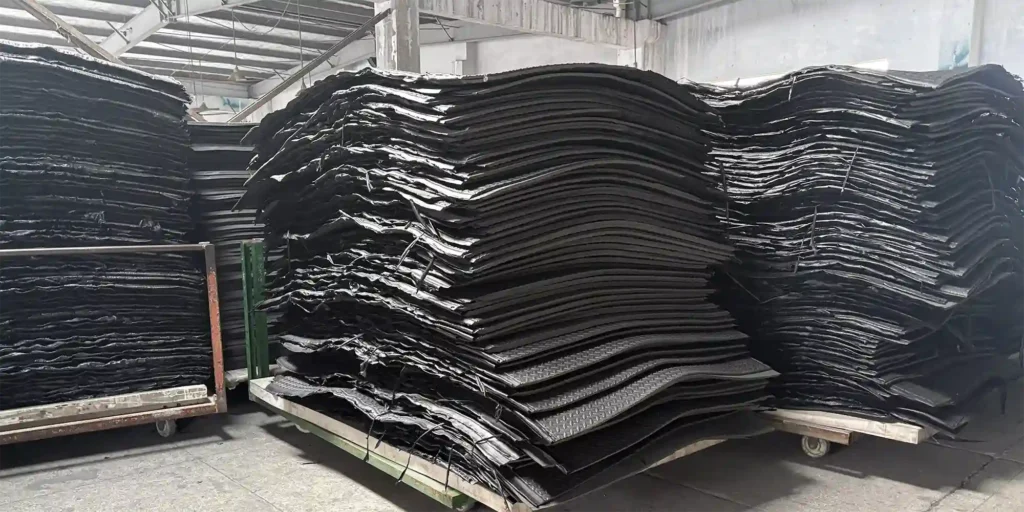 an indoor warehouse environment where several large stacks of black industrial sheets are piled on wheeled carts. The warehouse has a concrete floor and exposed metal beams in the ceiling. The sheets appear to be made of a sturdy material, likely intended for industrial or manufacturing applications. The scene indicates a functional storage area for these large components, with the carts allowing for easy movement within the facility.