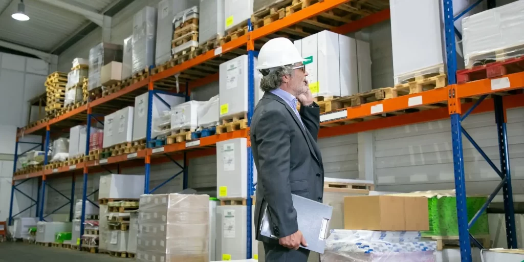A construction worker stands in a warehouse, laptop in hand. He is wearing a helmet and white coat and is checking the listings on the boxes. In the corner of the warehouse is a large crate that appears to be piled high. The worker is trying to locate the crate and verify that it is suitable for storage or transportation to its destination.