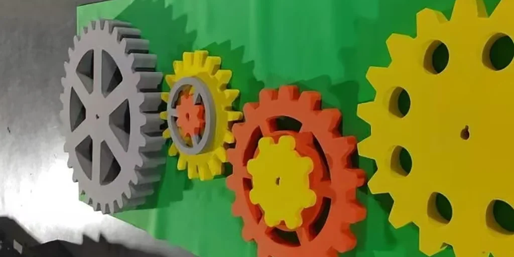 AlternativeText: “Colorful gears in gray, yellow, and orange mounted on a green background.” Caption: “A vibrant display of interlocking gears in gray, yellow, and orange on a green backdrop.” Description: “This image showcases a lively arrangement of gears affixed to a green surface. On the left, a large gray gear with a star - like design is visible. Next, a yellow gear contains a smaller orange gear at its core. Adjacent is an orange gear with a yellow gear layered on it. To the right, a large yellow gear with circular holes stands out. The mix of gray, yellow, and orange hues creates an engaging mechanical display, hinting at a playful or educational context.