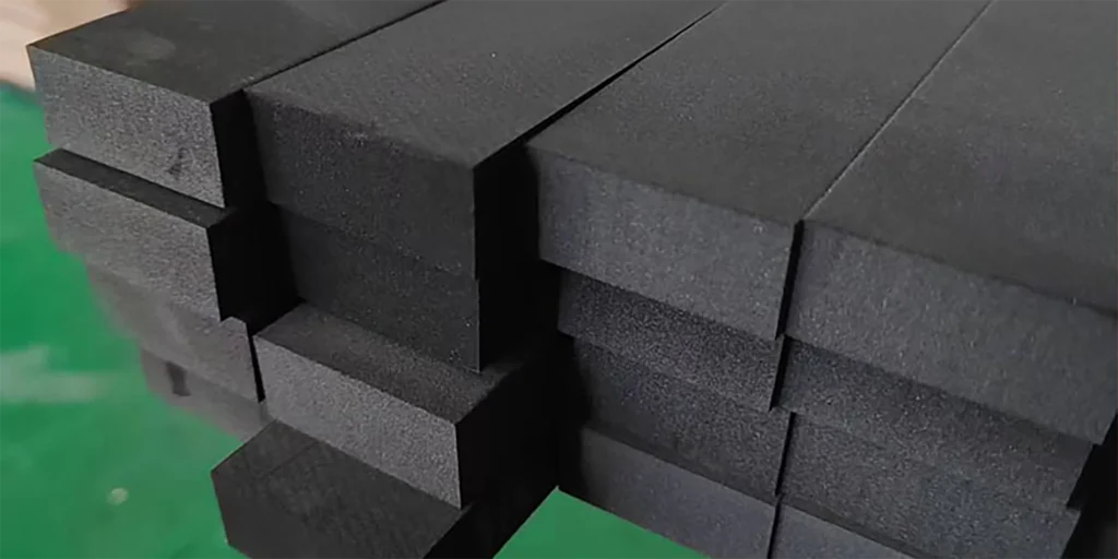 AlternativeText: Stack of black foam blocks with rectangular shapes. Caption: Stacked Black Foam Blocks with Precise Cuts Description: This image depicts a stack of black foam blocks, each cut into precise rectangular shapes. The blocks are arranged closely together, showcasing their uniform size and smooth surfaces. The green background provides a contrasting backdrop, highlighting the dark color of the foam. This setup suggests industrial or manufacturing use, emphasizing the precision and organization of the foam blocks.