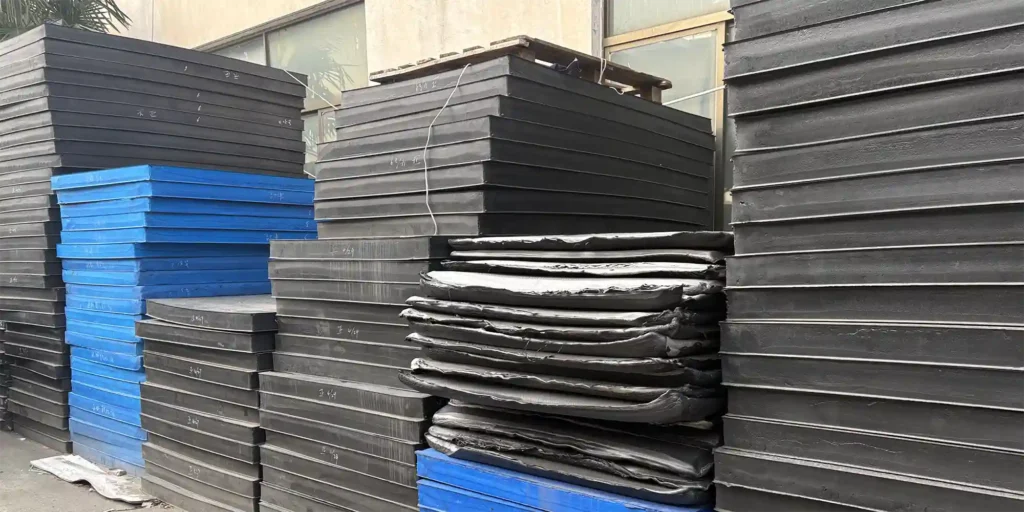 an outdoor industrial storage area where multiple large stacks of plates in black and blue are arranged. There is also a pile of black sheets among the stacks. The plates have handwritten markings, likely indicating specifications or inventory details. The scene includes a concrete ground, a building with windows, and palm trees, suggesting a functional storage environment for these large industrial components.