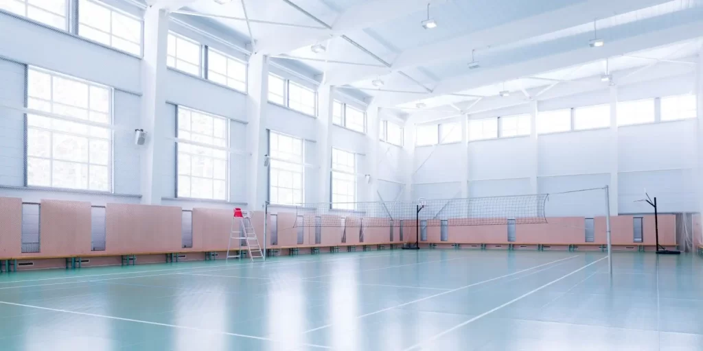 A spacious indoor gym with a huge window. It seemed to accommodate everyone, but there were only a few seats. A volleyball rack is placed in the center, while ball nets and badminton nets are located on both sides of the wall. Some sports equipment such as basketballs, soccer balls and volleyballs can be seen in the room. The whole space is covered by a green carpet to provide a cozy atmosphere.