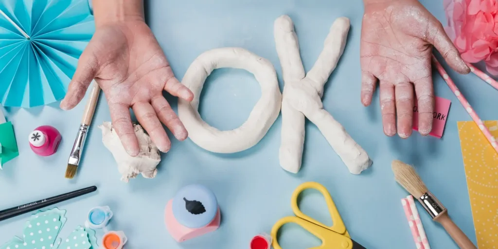 A scene from a craft production. On the tabletop, two hands are holding two white clay objects that have been molded to resemble letters. Various craft materials and tools are placed around the tabletop, such as colored paper flowers, scissors, glue, and brushes. These items seem to be prepared for some art project. The background is light blue, giving it a fresh and serene feel.