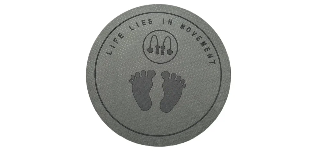 a circular emblem in a monochromatic gray tone. At the center, there are two symmetrical footprints facing each other, suggesting themes of balance and harmony. Above these footprints, there is a simple line drawing of a jump rope, indicating physical activity and exercise. Surrounding the central imagery, the phrase "LIFE LIES IN MOVEMENT" is inscribed along the circumference of the circle. The text is evenly spaced and written in capital letters, emphasizing the importance of motion and dynamism in life. The entire composition conveys a message about the value of staying active and engaged through movement.