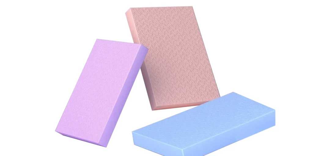 three rectangular blocks arranged against a white background. Each block has a distinct color: purple, pink, and blue. They appear to have a smooth, matte finish, resembling foam or sponge materials commonly used for crafting or packaging purposes. The blocks are positioned at different angles, creating a sense of depth and dimensionality. The lighting casts subtle shadows, enhancing their three-dimensional form. The overall composition gives a clean and minimalist aesthetic.