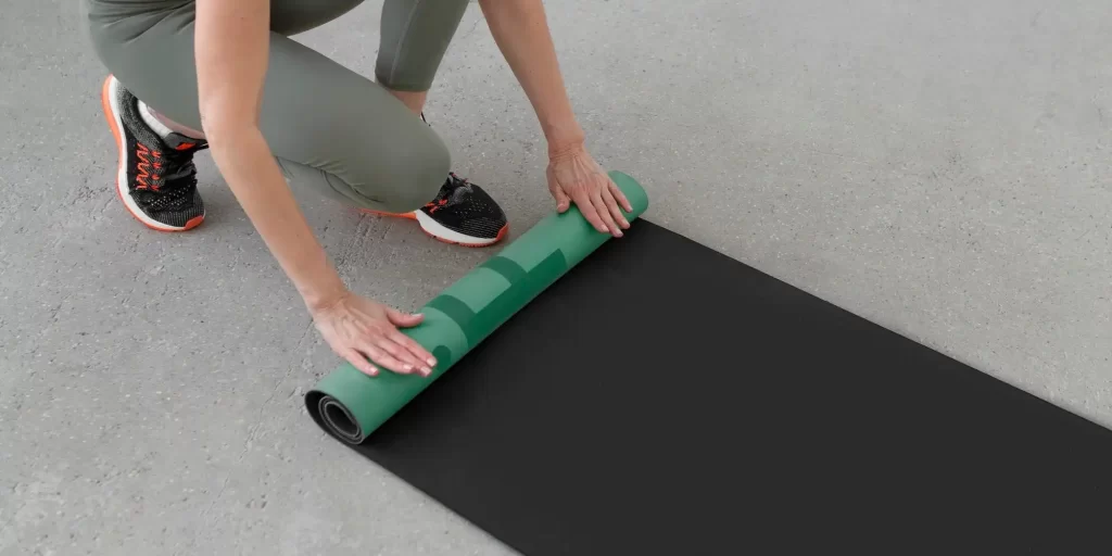Yoga mat, spread out on the floor, on which the woman is stepping to practice yoga. She was dressed in a green leotard and black pants, with her hair casually draped. Her hands were gripping the ends of the yoga mat and her body was leaning forward, as if she was searching for a sense of balance. Her focused and natural movements show the charm of yoga.