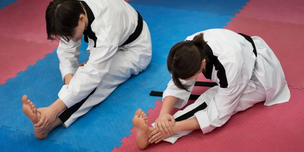 Two girls, dressed in white and black uniforms, standing on the floor or sitting on a bed with their feet crossed. They appear to be practicing kicking techniques. A boy stands behind another girl, a notebook in his hand, intently recording the details of the movements. They all seem to be very engaged and are working hard to train their skills. The scene is full of energy and passion, reminding people of those sweating it out in the gym.