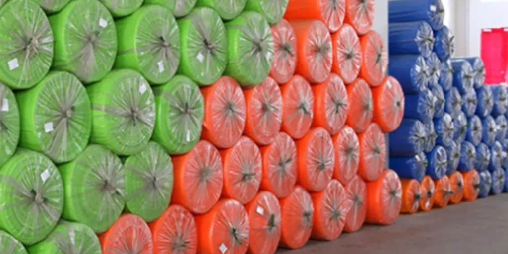 depicts an indoor warehouse storage area where large quantities of rolled materials are stacked. The rolls (wrapped in clear plastic) are primarily divided into bright green and orange sections, with additional blue rolls visible in the background. The neat, organized piles suggest these are industrial or manufacturing materials stored for distribution or production use, typical of a large-scale storage facility.