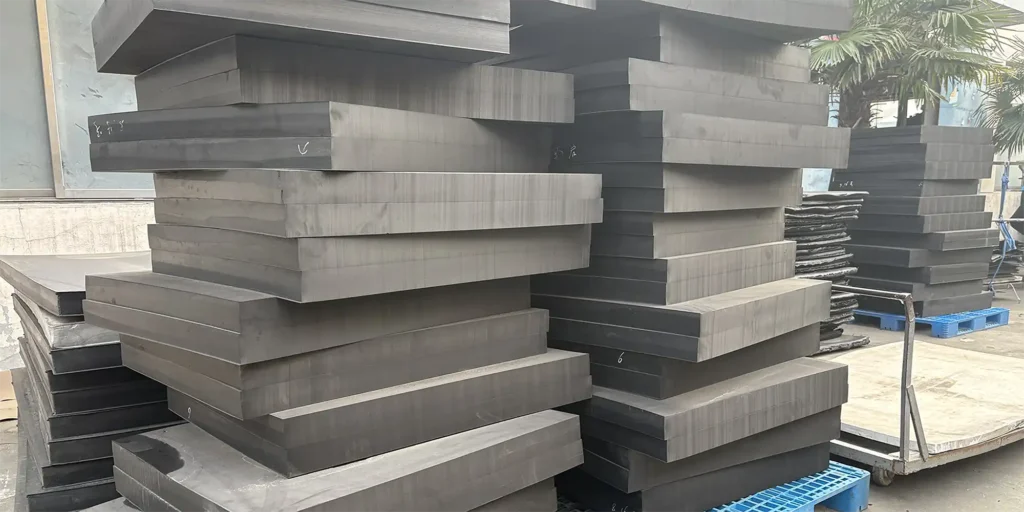 an outdoor industrial storage area where massive stacks of thick black industrial blocks are arranged on pallets. The blocks appear to be made of a sturdy material, likely for heavy - duty industrial or manufacturing use. The scene includes a concrete ground, a building with windows, palm trees, and other industrial storage elements like additional stacks and a cart. The presence of markings on some blocks suggests inventory or specification details, indicating a functional storage environment for these large components.