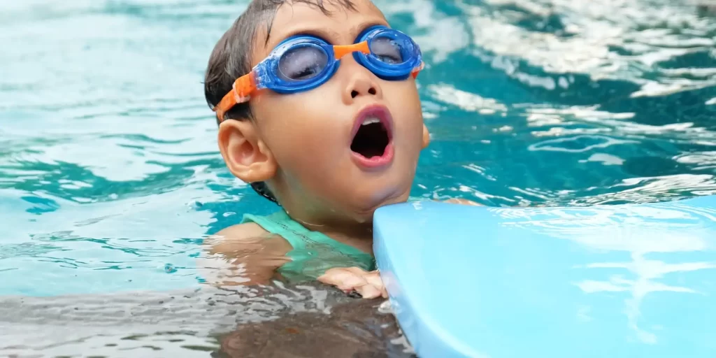 The child is swimming in the water, wearing goggles and glasses. His mouth is open and he seems to feel excitement or surprise. This is a delightful scene that shows the child's joy and vigor. Children can feel relaxed and free when they play in the pool, which makes it more enjoyable for them.