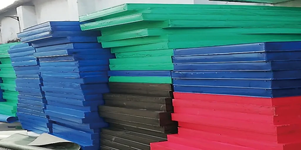 depicts an outdoor industrial storage scene where multiple large stacks of plates in bright colors—green, blue, black, and red—are arranged. The plates are neatly piled, and the background includes parts of a building, indicating a functional storage environment for these colorful industrial components. The variety of hues suggests different material types or specifications for manufacturing use.