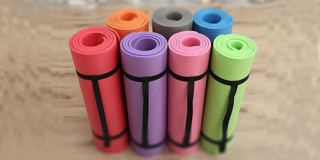 six different colors of yoga mats placed on a wooden floor. From left to right, the colors are red, purple, pink, gray, blue and green. Each yoga mat is rolled up and has a black carrying strap attached to it. These colorful yoga mats provide a variety of choices for users according to their preferences.