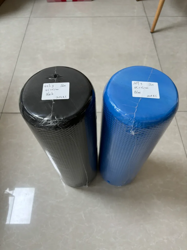 This image shows two cylindrical EVA yoga poles that have been placed on a light colored tile floor. 1. the EVA yoga pole on the left is black and has a white label affixed to it with text that shows the color, weight, size, and date of the object. 2. The EVA yoga pole on the right is blue and also has a white label with the same information as the label on the black EVA yoga pole.