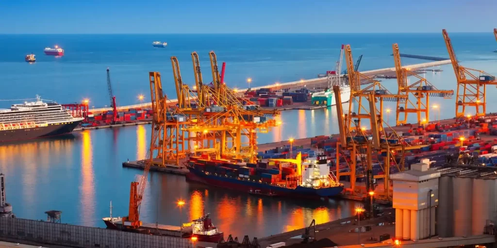 A port city, mesmerized by the ships and lights intertwined at night; the bow of a ship towering as huge as a giant ship, docked at the wharf. Large cranes and hoists work busily to provide the necessary support for the ships. There are also numerous containers in the harbor, waiting to be loaded or unloaded with cargo. The beauty of the harbor at night is mesmerizing, as if you were in a dream.